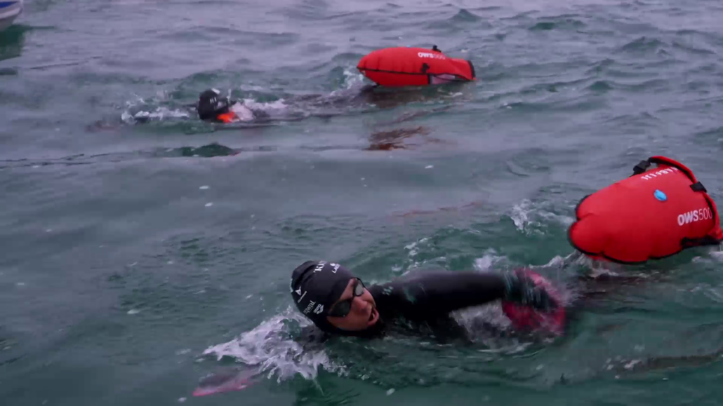 Two swimmers in wetsuits are swimming in open water with red buoy floats attached to them. They are surrounded by choppy waves, and one swimmer is in the foreground while the other is slightly behind. The scene appears to be part of The Madiba Challenge, an intense open-water race or training session.