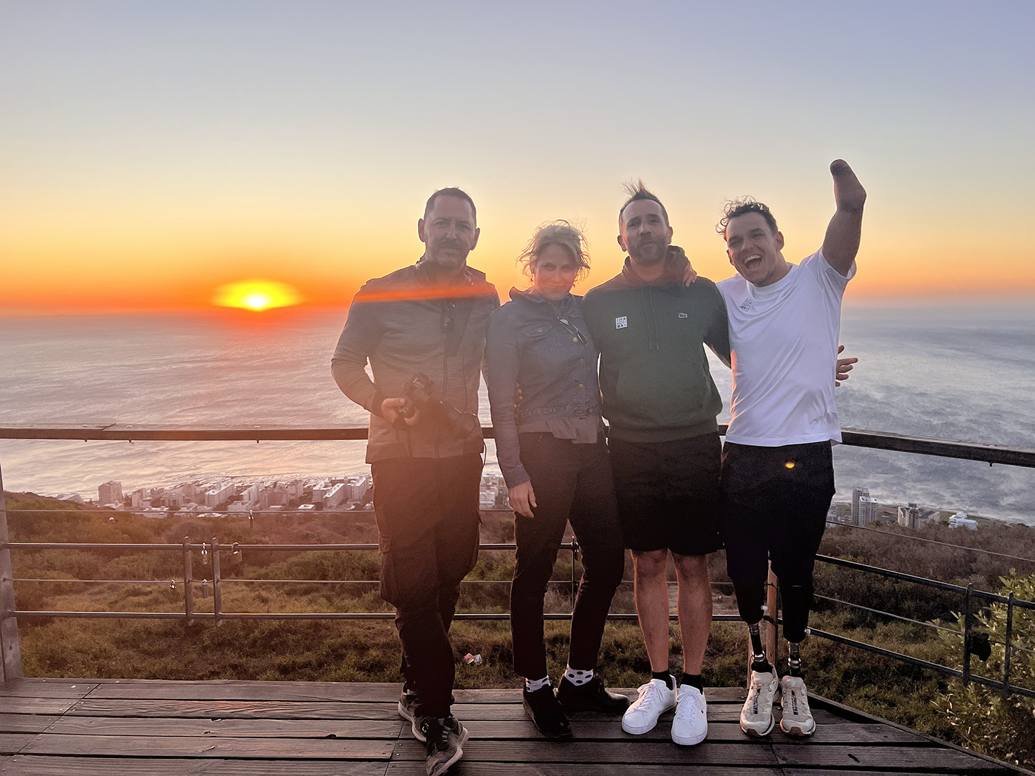 Four people stand together on a wooden deck overlooking an ocean sunset. The group, dressed in casual outdoor clothing, is smiling and posing for the photo, with two men on each side and a woman and another man in the center. Having completed The Madiba Challenge, they savor the scenic coastline below them.