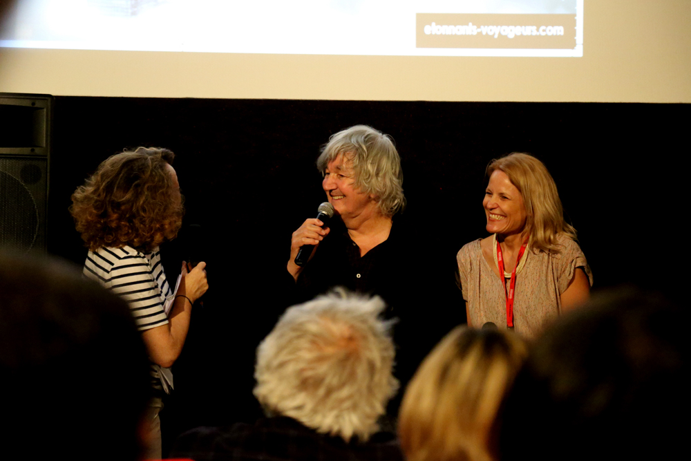 Three people are on stage at an event. The person in the middle is holding a microphone and speaking, recounting a shaman’s journey, while the other two look on and smile. The person on the left has curly hair and is wearing a striped shirt. The person on the right has straight hair and is wearing a red lanyard.