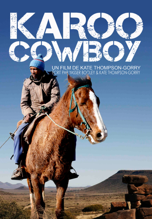 A man in rugged clothing rides a brown and white horse through a mountainous desert landscape under a clear blue sky. "Karoo Cowboy" and credits appear at the top of the image, perfectly encapsulating the spirit of Western adventure.