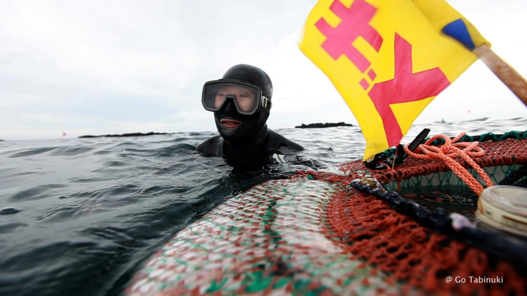 A person wearing a black diving suit and goggles is partially submerged in the water near a colorful fishing net. A yellow and red flag with a symbol waves from the net. The sky is overcast, and the ocean surface is slightly choppy. © Go Tabinuki.