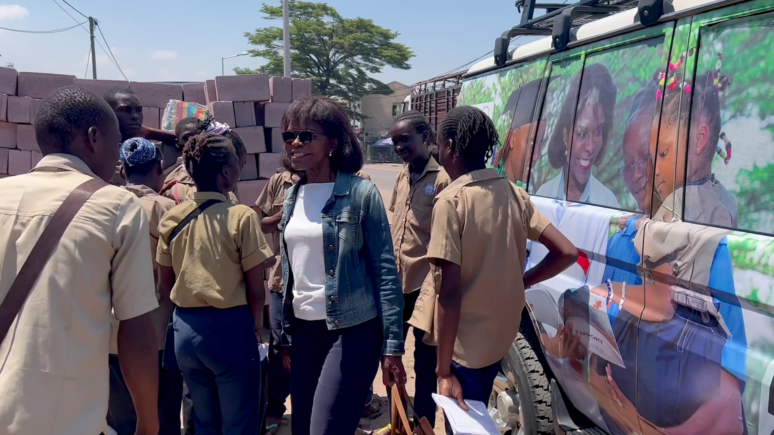 A woman in sunglasses and a denim jacket, identified as Francine, stands next to a group of young students in uniforms. They are gathered around a vehicle with large photos of children on its side. The scene appears to be outdoors on a sunny day, showcasing Francine’s African ambition for children's education.