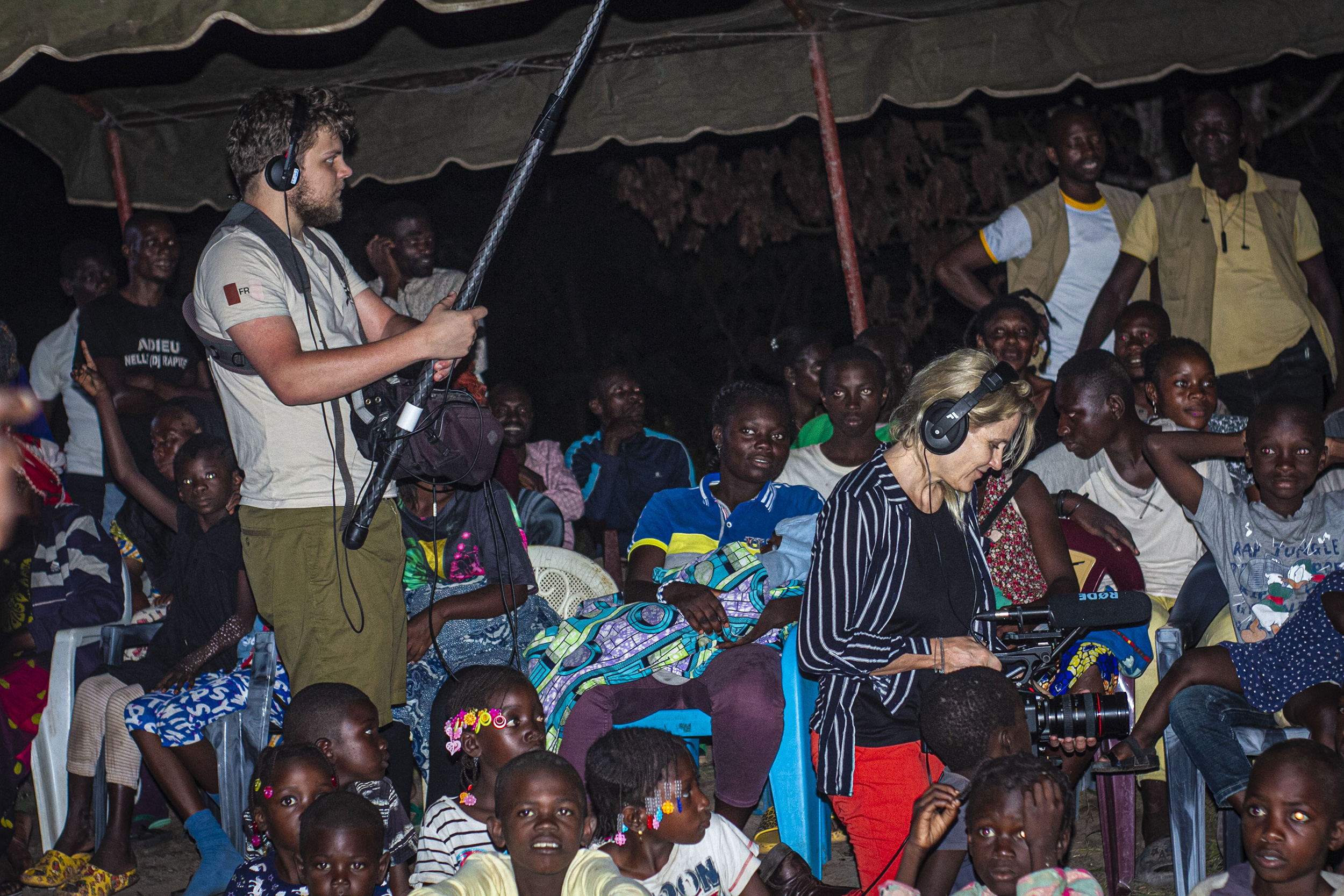 A group of people, including children, gather outdoors at night, some sitting under a canopy. Francine, an African filmmaker with boundless ambition, is in the foreground with her team; one holds a boom mic while another operates a camera. The atmosphere is lively and engaged.