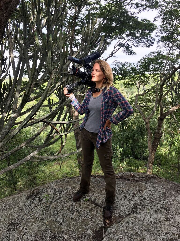 A woman stands on a large rock holding a video camera. She wears a plaid shirt over a gray top, green cargo pants, and brown boots. She is surrounded by lush greenery with various trees in the background. The sky is partly cloudy.