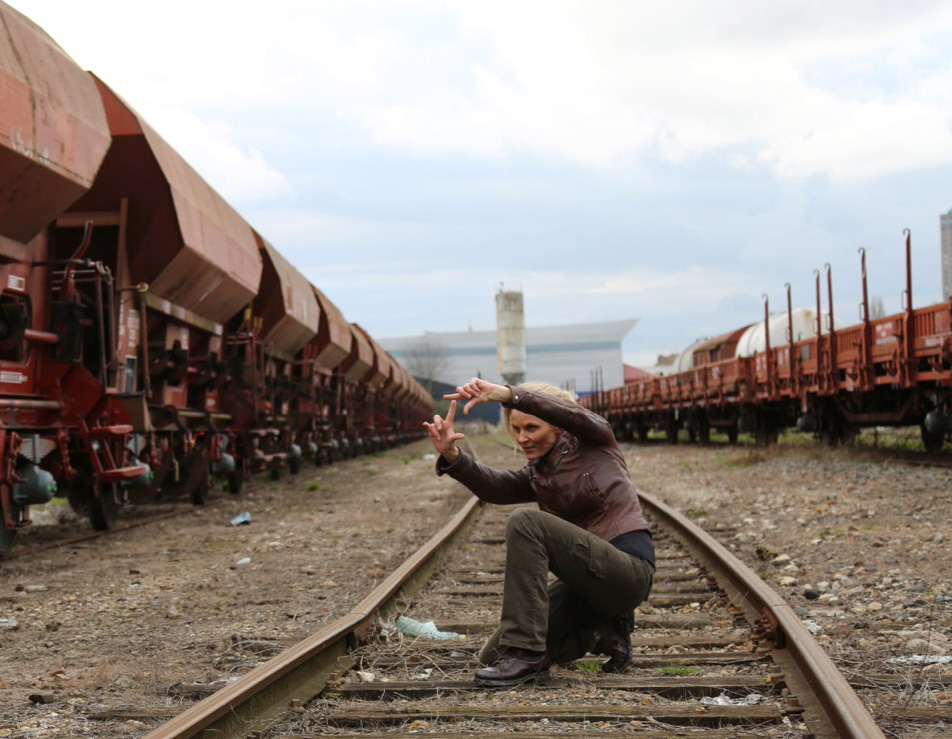 A person squats on railway tracks, surrounded by rows of orange freight cars on both sides. They have short hair and wear a brown jacket and green pants. The sky is cloudy, creating an industrial backdrop.